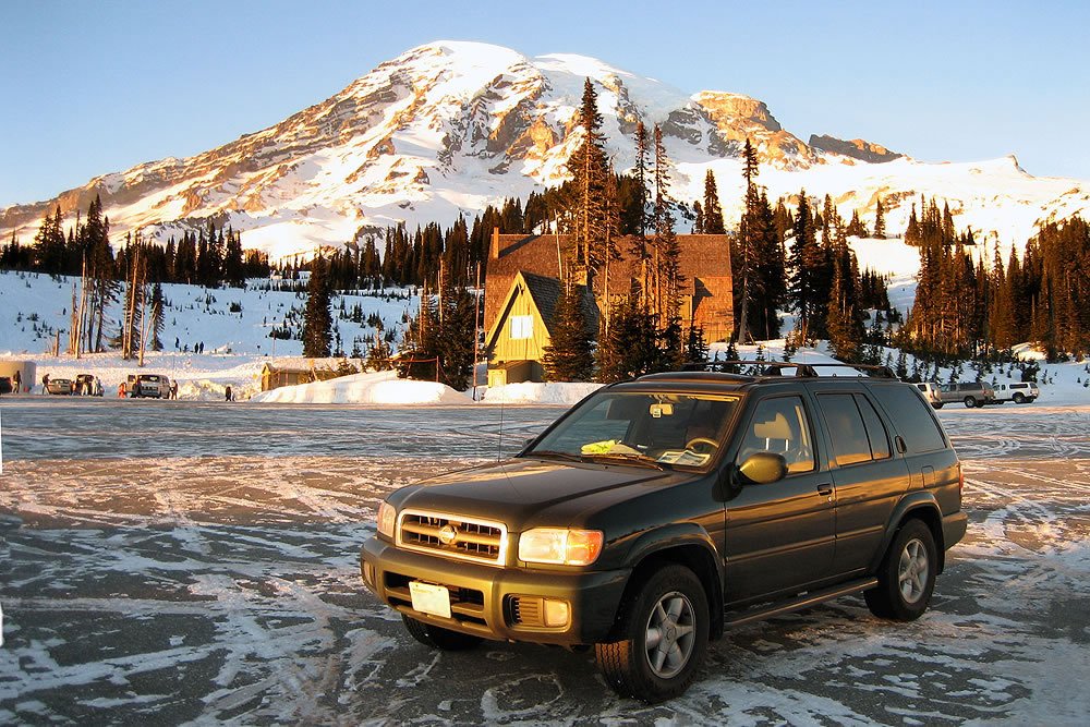 A green Jeep Wrangler parked on icy terrain in front of a snow-covered mountain lodge under clear blue skies.