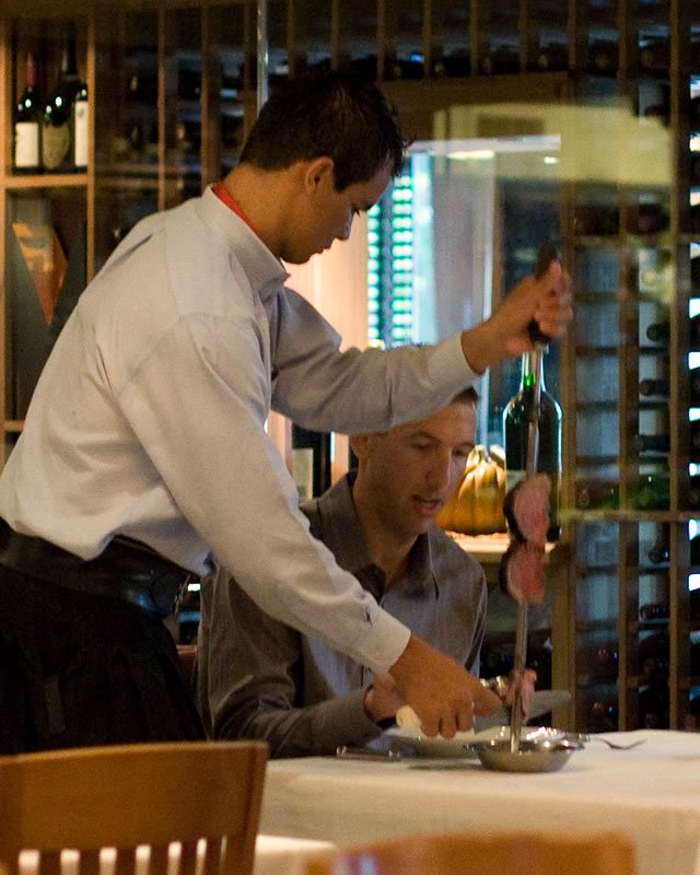 John is pouring wine from a bottle into a glass while Kristie looks on. They are seated at a table with wine bottles in the background.