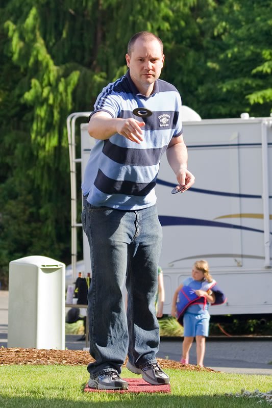 A man in a striped shirt and jeans is focused on throwing a disc at a shuffleboard court, with lush green trees and a child in the background.