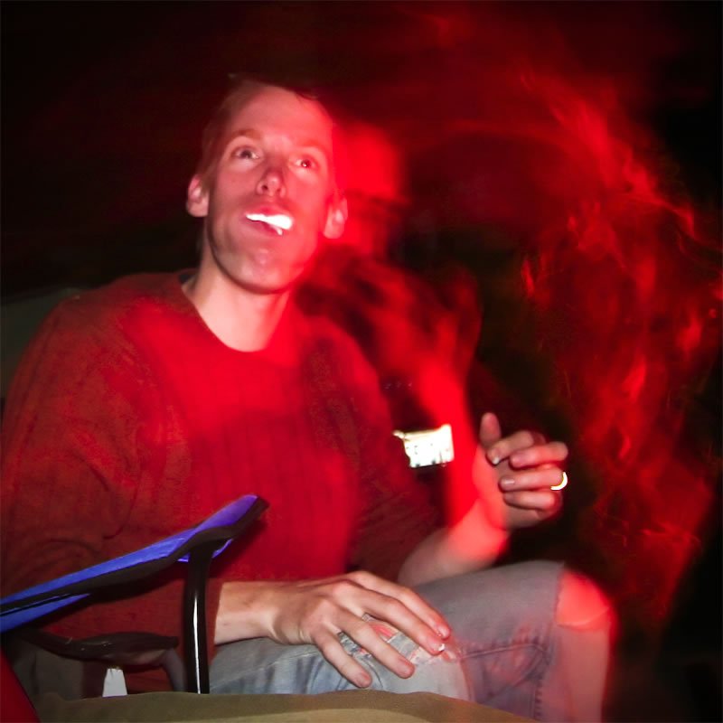 John sits smiling under red lighting, holding a cup and wearing a red sweater.