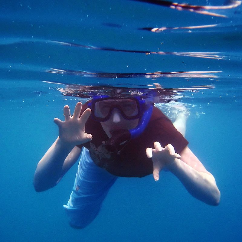 John is captured underwater while snorkeling, wearing a mask and gesturing with his hands in clear blue water.