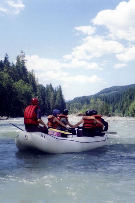 A group of friends in a raft down a river surrounded by forested hills under a blue sky with clouds.