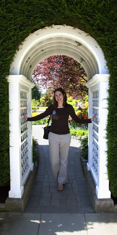 A woman walks through a white arched gate in the garden, surrounded by lush greenery and a vibrant red-leafed tree.