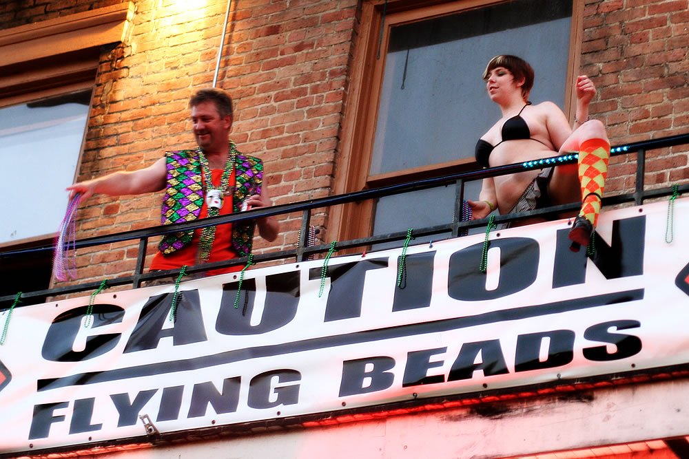 Couple throwing beads on a street parade with a sign warning caution flying beads.