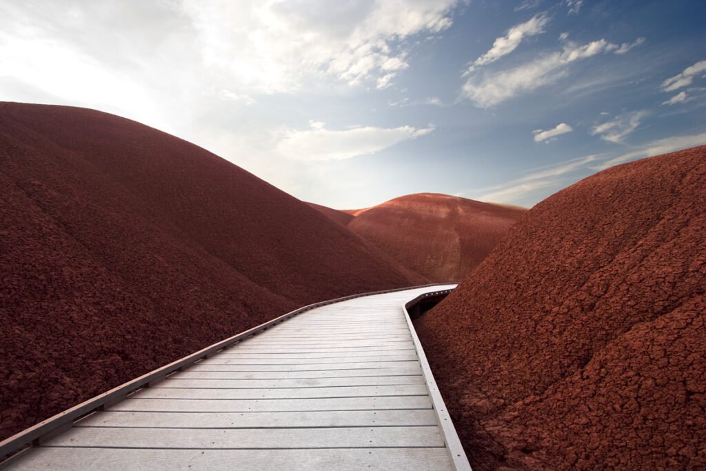 A wooden walkway winds through vibrant red dunes under a blue sky with fluffy clouds.