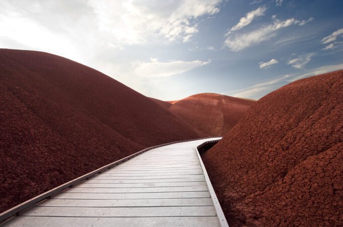 A wooden walkway winds through vibrant red dunes under a blue sky with fluffy clouds.