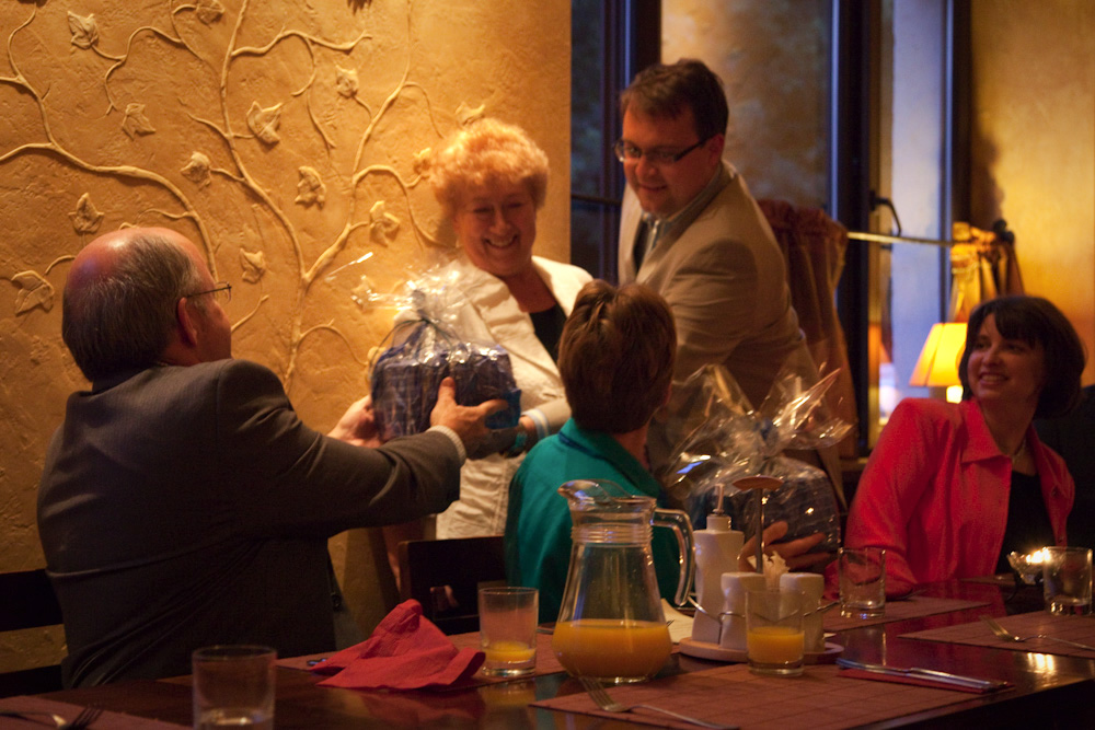 A group of people sitting around a dinner table, sharing laughter and holding gifts. The setting features warm lighting and decorative wall art.