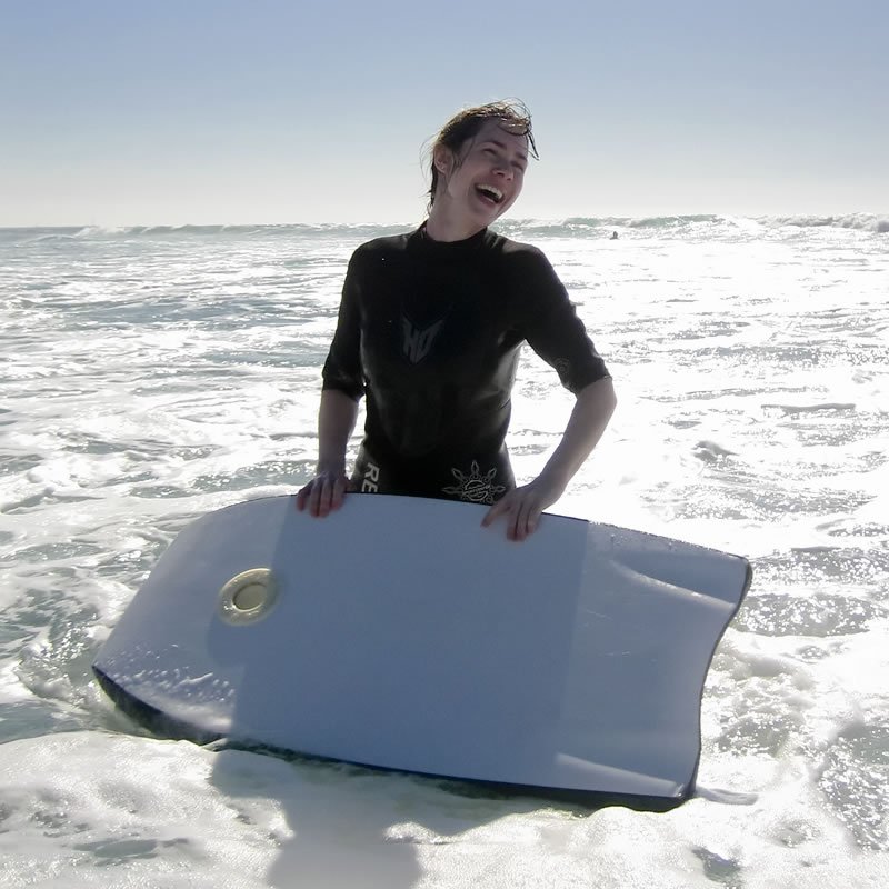 A smiling person in a wetsuit stands in the ocean holding a surfboard, capturing a moment of happiness at the beach.