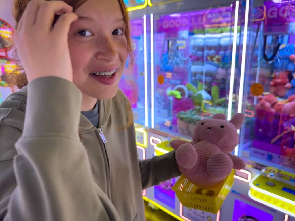 A person smiles while holding a freshly won stuffed toy from a claw machine, surrounded by the colorful lights of an arcade setting.
