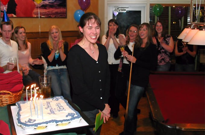 A woman stands in front of a lit birthday cake surrounded by a smiling and clapping group of people, festive balloons in the background.