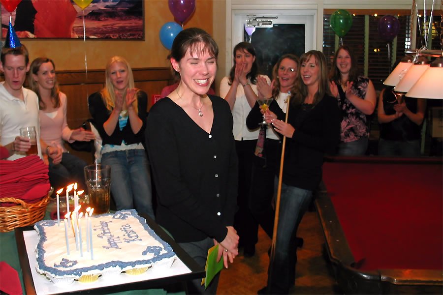 A woman stands in front of a lit birthday cake surrounded by a smiling and clapping group of people, festive balloons in the background.