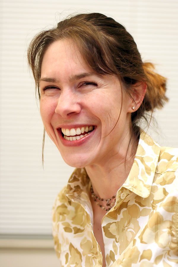 A woman with brown hair styled in a bun, wearing a yellow patterned blouse, laughing with visible joy.