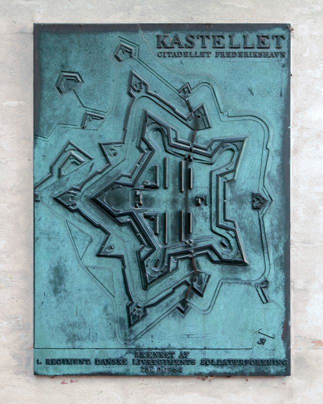 Close-up view of a weathered bronze plaque showing a relief map of the Kastellet fortress in Copenhagen. The metallic sign details the star-shaped layout of the historic military citadel on Christianshavn.