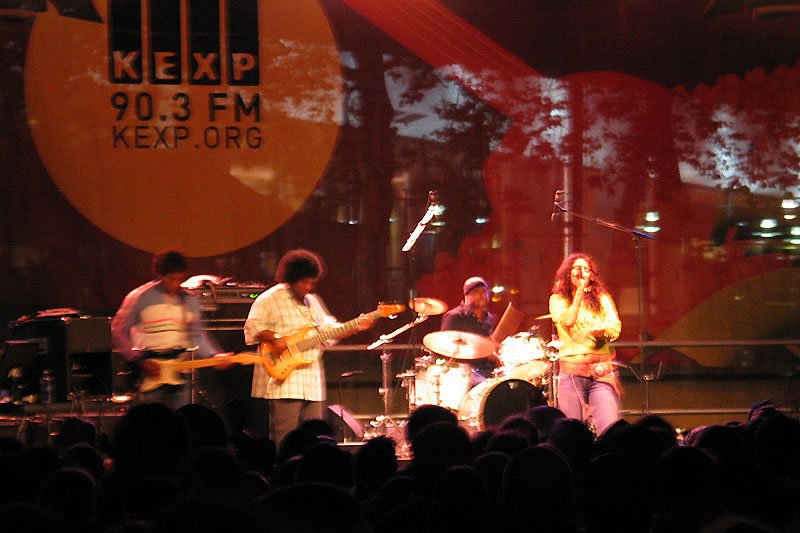 A live concert performance on stage with KEXP 90.3 FM branding, featuring musicians playing instruments under warm stage lighting, with an audience silhouetted in the foreground.