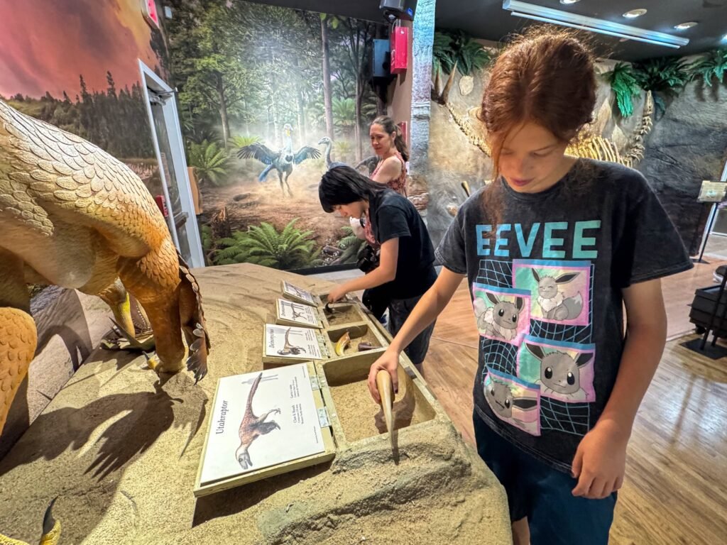 Children dig in sand bins and examine dinosaur models at a museum exhibit designed to simulate paleontological exploration.