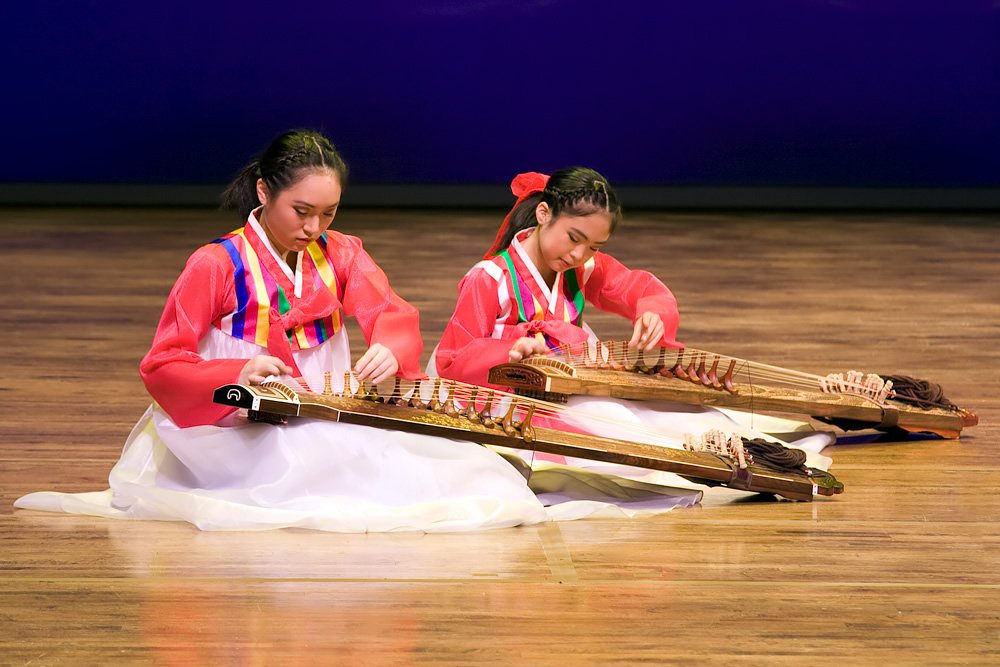 Two performers in vibrant traditional Korean hanbok seated on stage, skillfully playing the gayageum. The background features a deep blue curtain, highlighting the rich colors of their outfits.