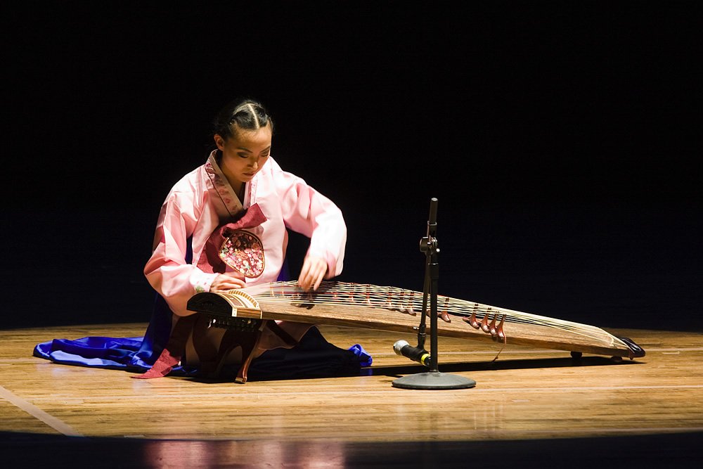 A young girl in traditional attire plays a gayageum on stage.