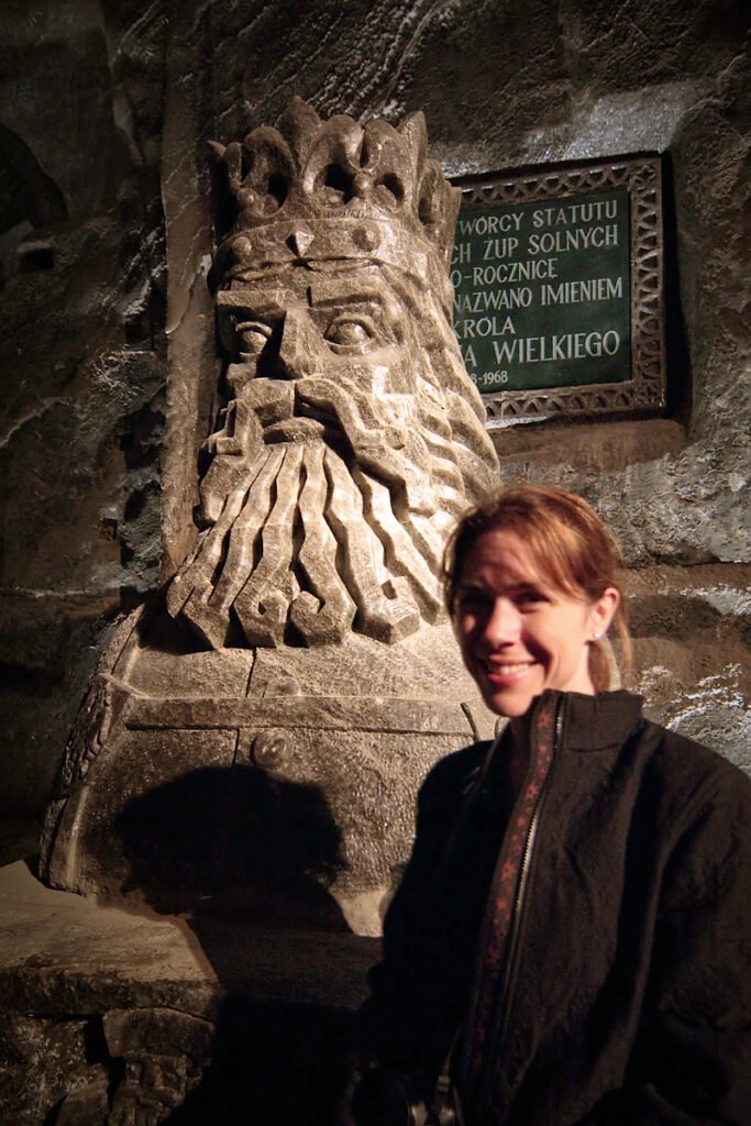 Kristie smiles next to a stone statue of King Arthur, with a plaque behind displaying Polish text.