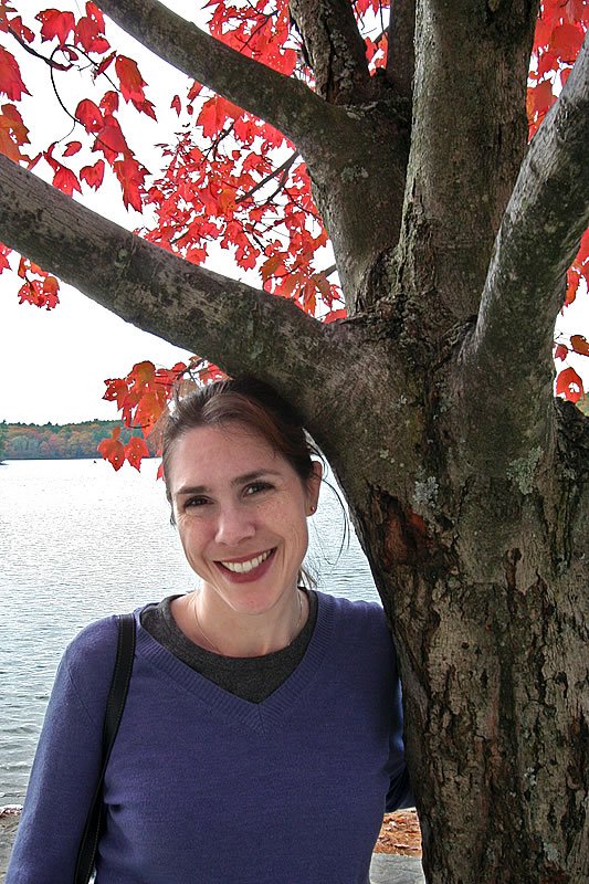 Kristie smiles warmly beside a tree with vibrant red autumn leaves overlooking water in the Pacific Northwest.