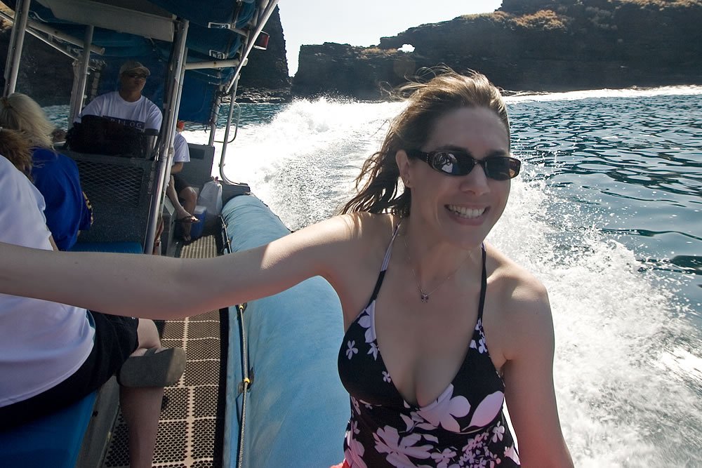A smiling woman in a floral swimsuit and sunglasses leans out from a moving boat with ocean spray behind her.