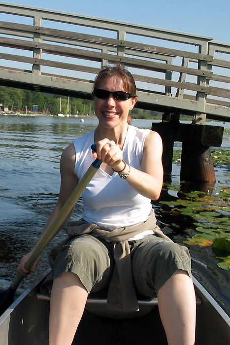 Kristie smiling in a canoe while paddling under a rustic wooden bridge on a calm lake surrounded by lily pads and greenery.