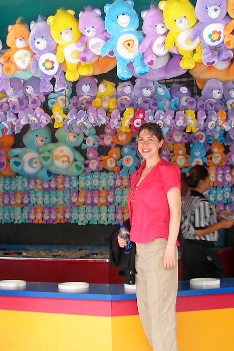 Kristie in a pink shirt smiles in front of a carnival game stacked high with colorful Care Bears plushies. The background shows a vibrant game booth filled with these nostalgic teddy bears.