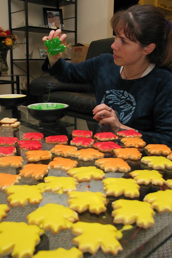 Kristie decorates fall-themed cookies with green icing in a cozy kitchen. The scene shows festive, leaf-shaped cookies in vibrant colors ready for the holiday season.