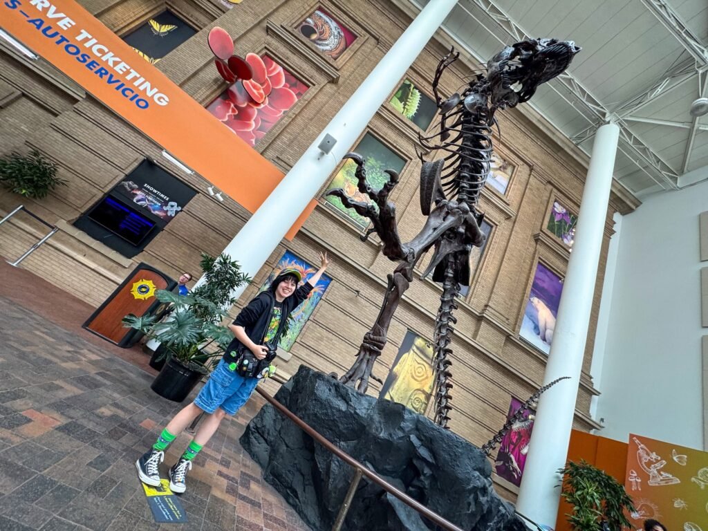 A woman wearing blue shorts and bright green socks stands next to a large dinosaur skeleton exhibit. She is posing near a white pillar inside a museum hall.
