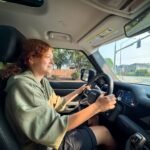 Kristie with curly red hair is driving a car, wearing a green hoodie and black shorts.