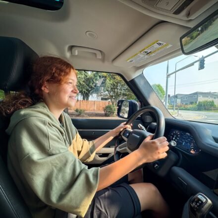 Kristie with curly red hair is driving a car, wearing a green hoodie and black shorts.