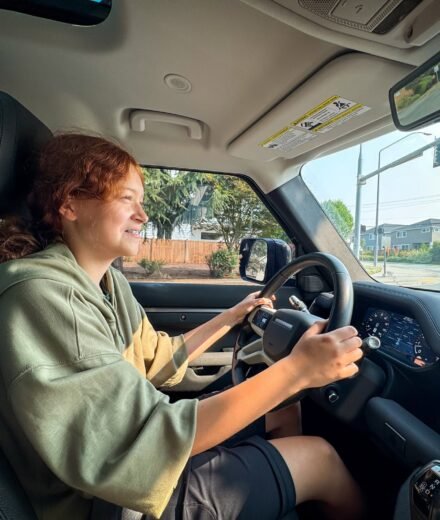 Kristie with curly red hair is driving a car, wearing a green hoodie and black shorts.