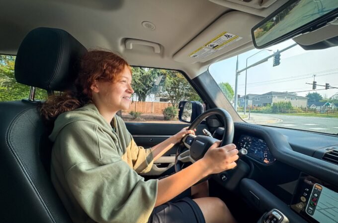 Kristie with curly red hair is driving a car, wearing a green hoodie and black shorts.