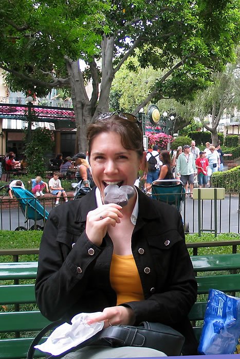 Kristie is seated on a green park bench, delighted as she enjoys ice cream. The background shows a lively park with people and lush greenery.