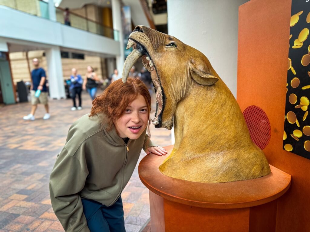A woman smiling next to a sculpted lion head inside a modern museum space. She leans her head close to the lion’s open mouth in a playful pose.