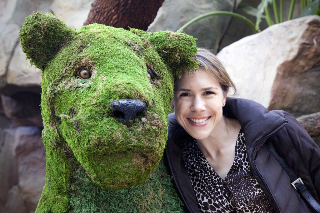 A woman smiles next to a giant green moss-covered polar bear figure in a zoo setting