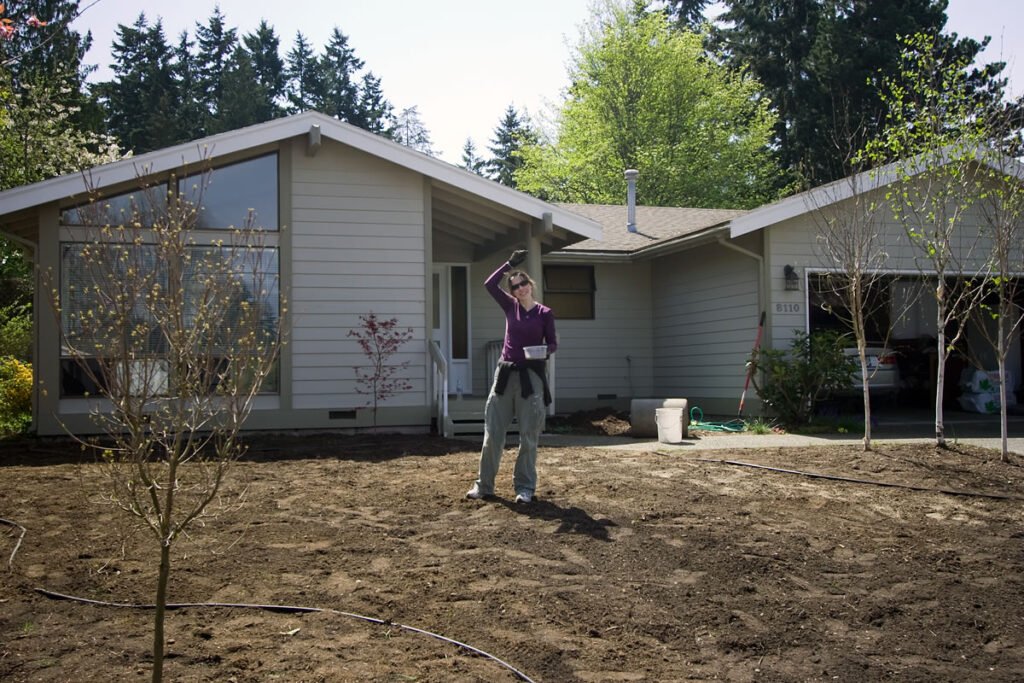 A woman stands in a dirt yard in front of a beige house with green trees in the background.