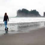 Kristie walks alone on a wet, sandy beach on an overcast day. Misty sea stacks are visible in the distance across the ocean.