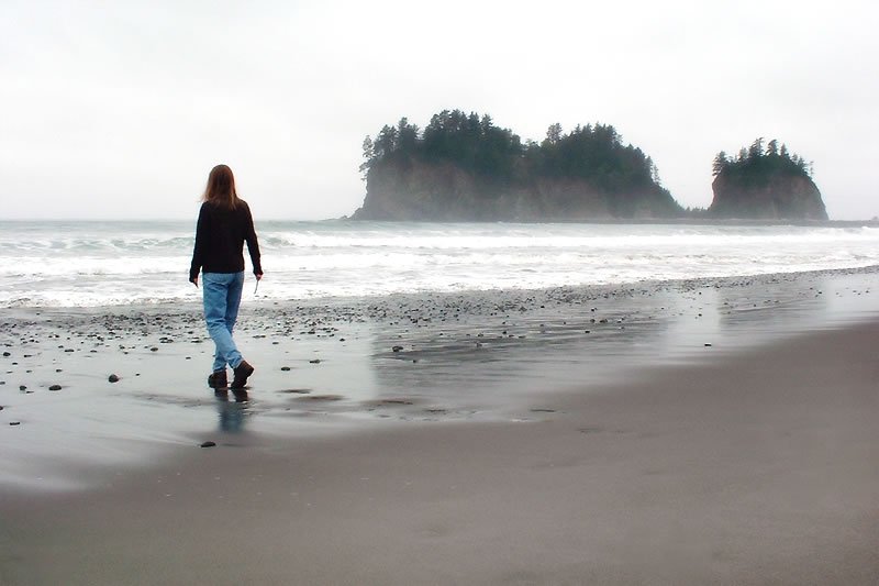 Kristie walks alone on a wet, sandy beach on an overcast day. Misty sea stacks are visible in the distance across the ocean.