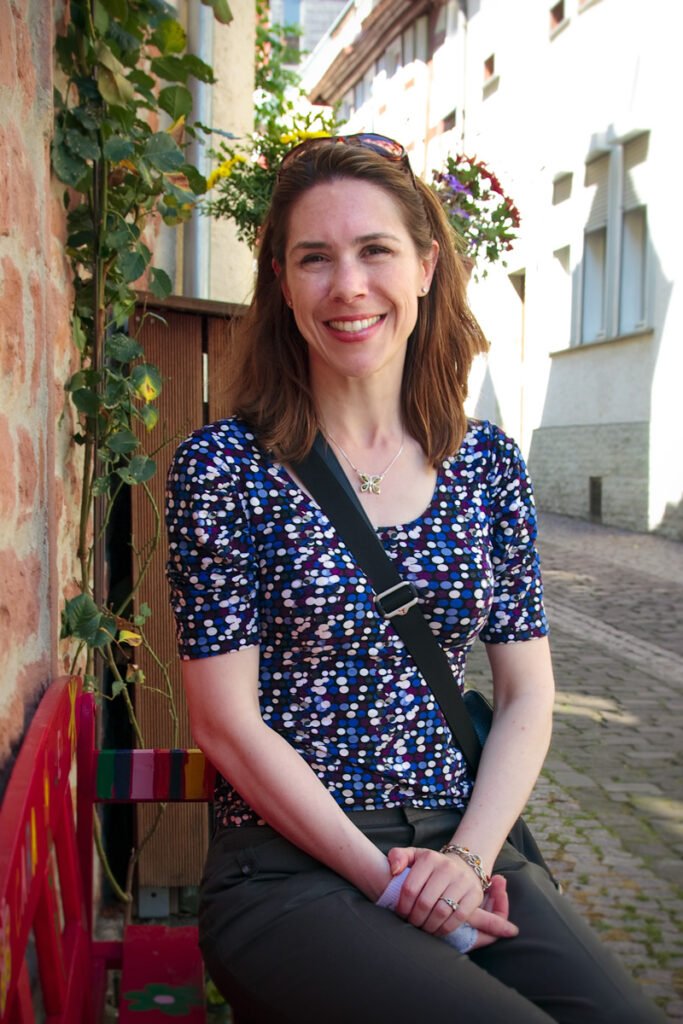 A smiling woman sits on a red bench outdoors near a brick wall adorned with vines and flowers.