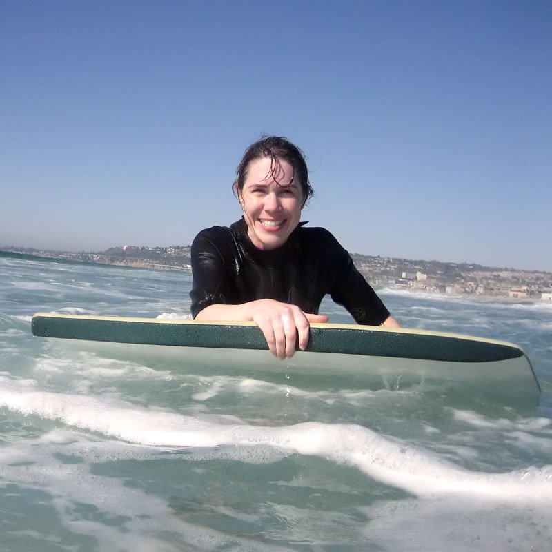 Kristie in a wetsuit paddling on a surfboard with a scenic coastal backdrop. Blue skies and ocean waves surround her.