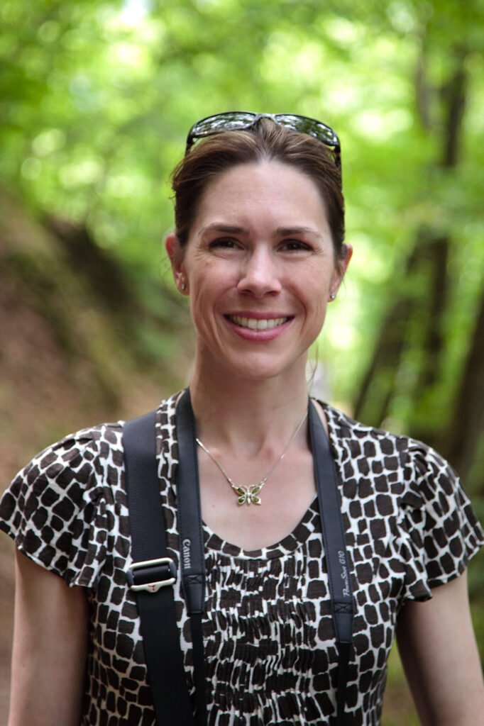 A smiling woman wearing a patterned top and sunglasses on her head, standing in front of a lush green forest background.