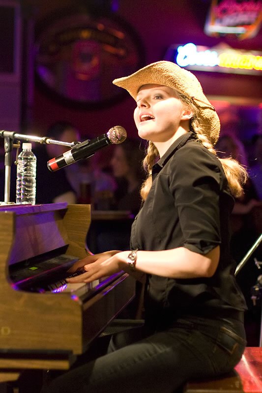 Kristie sits at a piano, singing into a microphone while wearing a beige hat and black outfit. The background shows vibrant stage lighting with purple and blue hues.