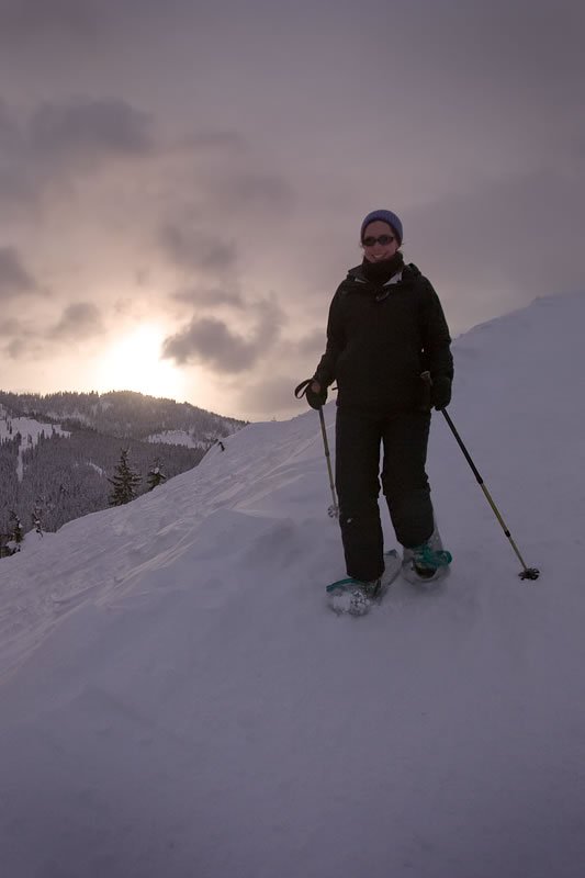 Kristie stands on a snowy mountain slope with ski poles, the sun setting behind her.