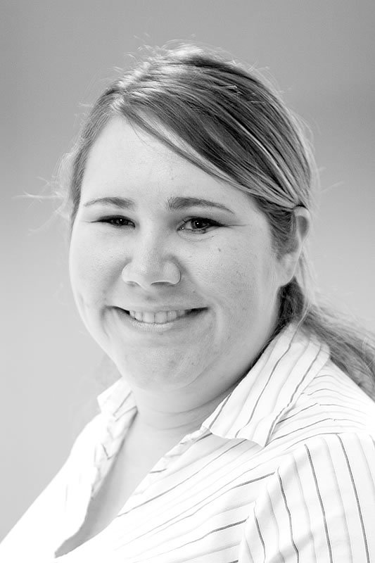 A black-and-white photo of Kristie smiling warmly, wearing a striped collared shirt with her hair pulled back.