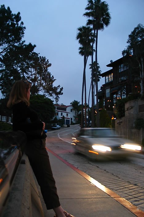 A woman stands on a sidewalk in Los Angeles at dusk. A car's headlights blur past as palm trees rise against the twilight sky.