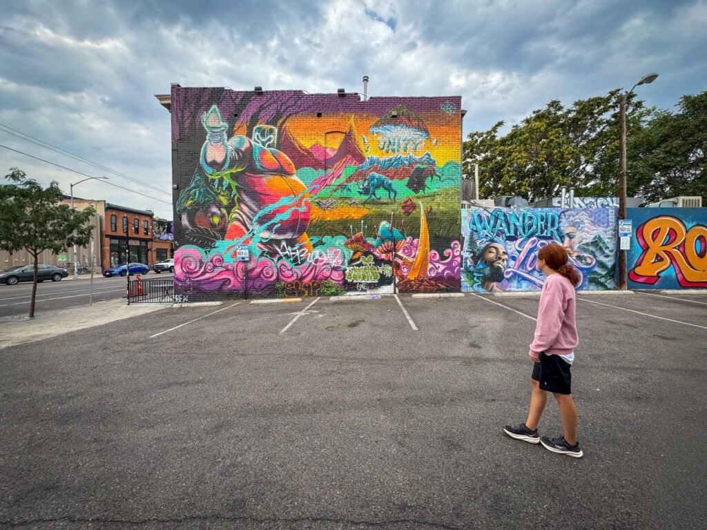 A woman in a pink hoodie walks through a parking lot toward a colorful graffiti-covered wall under a cloudy sky.