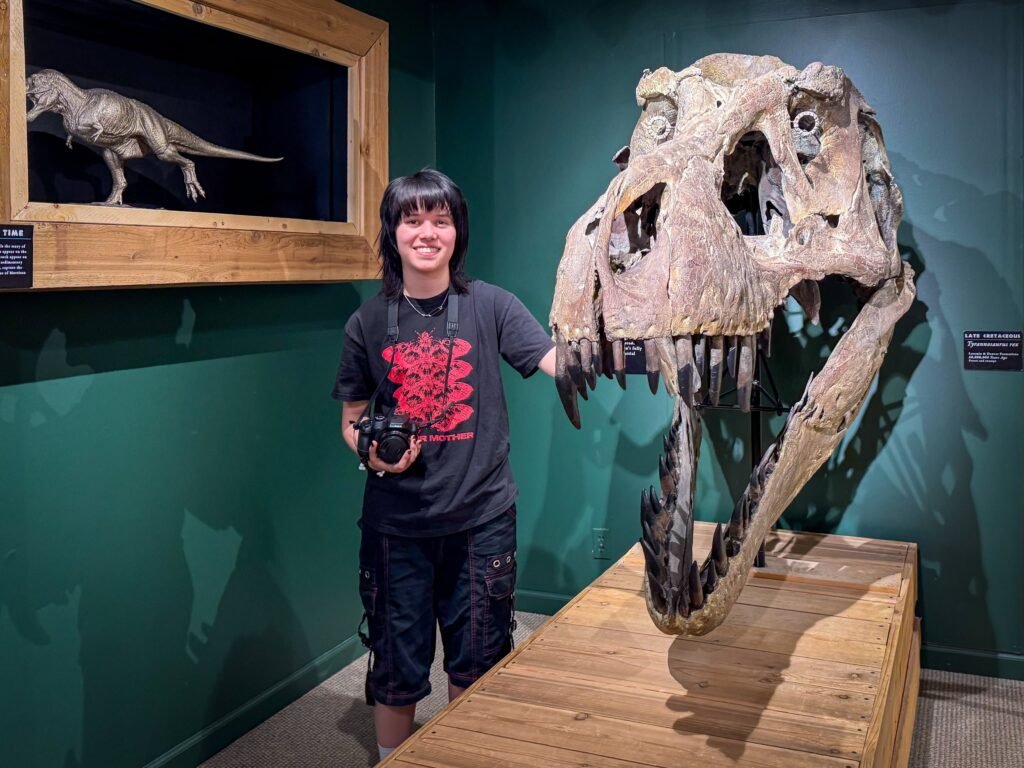 A smiling person stands beside a large T-Rex skull fossil on display, with a dinosaur model in a glass case behind them.