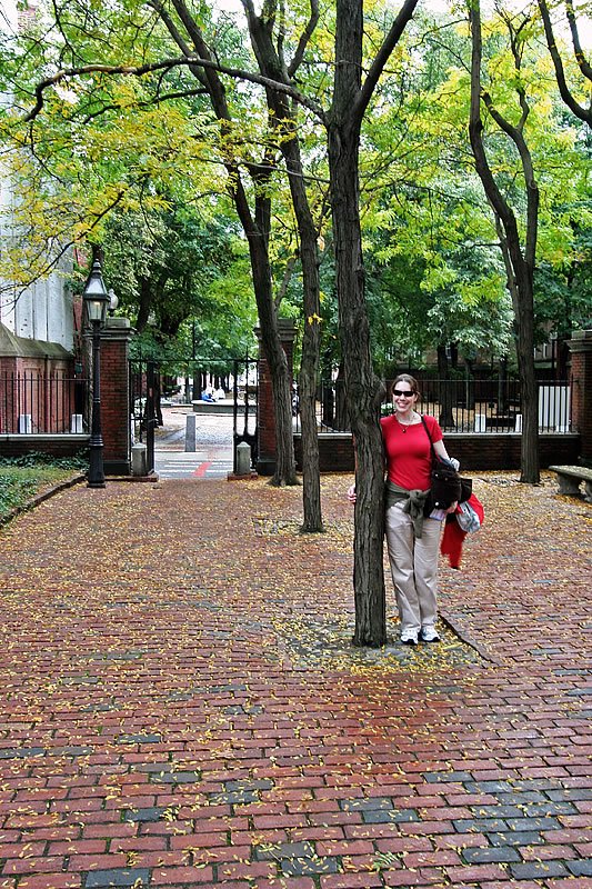 A woman in a red shirt stands by a tree holding a black bag on a brick pathway.