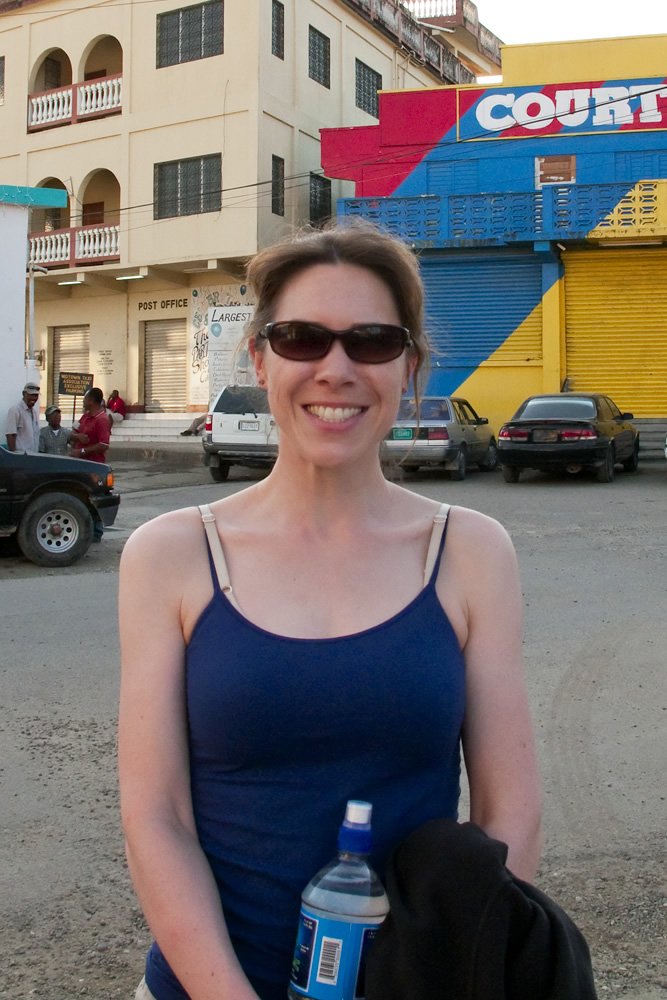 Kristie wearing sunglasses and a blue tank top, holding a water bottle and smiling in front of a colorful building with 'COURT' written on it.