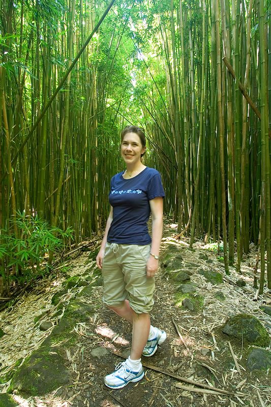A woman wearing a blue shirt and khaki shorts stands smiling in a bamboo forest. The dense greenery surrounds her as she enjoys the natural setting.